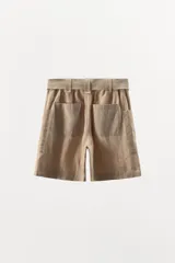 Bermuda color beige de lino, con cintura elástica, trabillas y cinturón con hebilla metálica. Presenta bolsillos de parche laterales y traseros.