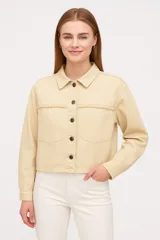Sobrecamisa color beige de corte cropped, con cuello camisero, cierre frontal con botones a tono, mangas largas y dos bolsillos con solapa y flecos en el pecho.