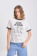 Remera color crudo de manga corta con cuello y puños negros. Estampa frontal con texto "Côte D'Azur" y dibujo de playa con palmeras.