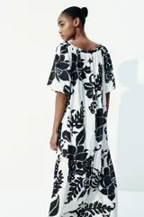 Vestido largo de corte holgado con estampado floral en blanco y negro. Tiene escote cuadrado con cordón ajustable y mangas cortas abullonadas con elástico en los puños. Presenta costura fruncida bajo la cadera.