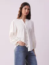Blusa blanca de gasa con cuello a la base, escote en V y abertura central con botones. Tiene mangas 3/4 amplias con apliques de crochet y detalles plisados en la parte delantera.