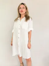 Vestido camisero corto de lino blanco, con cuello clásico, cierre frontal con botones, manga corta con puños y bolsillo de parche en el pecho. Corte holgado y bajo redondeado.