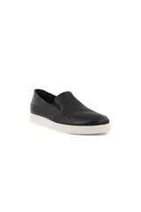 Zapatilla tipo slip-on de cuero negro, con suela blanca gruesa. La parte superior presenta un patrón de perforaciones en forma de ondas o líneas diagonales.