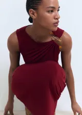 Vestido midi color borgoña, de corte ajustado, sin mangas y con detalle drapeado en el lateral. Presenta un adorno dorado en el hombro.