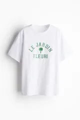 Remera blanca de algodón con corte amplio, cuello redondo y mangas cortas. Estampado frontal con texto en francés "Le Jardin Fleuri" y una palmera.