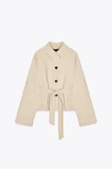 Trench corto color crema con cuello solapa, manga larga con trabilla, bolsillos de doble vivo, forro interior y cinturón lazada en el mismo tejido.