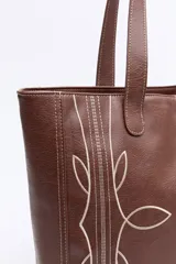 Cartera tipo tote bag en símil cuero color marrón oscuro, con bordados decorativos en blanco que evocan un estilo western. Cuenta con asas largas para llevar al hombro.