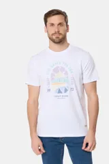 Remera blanca de manga corta con cuello redondo y un estampado gráfico frontal que simula un paisaje costero con un sol naciente o poniente, rodeado por texto circular que dice "SURF - LIFE UNDER THE SUN - CLUB".