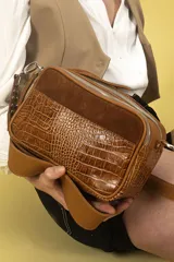 Cartera bandolera de cuero color marrón con estampado croco y detalles de gamuza color óxido. Tiene correa ancha regulable con tachas metálicas y doble cierre superior independiente.