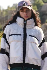 Campera de fleece de pelo alto color blanco, con cuello alto, cierre frontal completo y detalles en negro en el cierre, puños, cintura y franjas en las mangas. Incluye bolsillos frontales con solapa en color blanco liso.