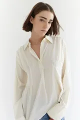 Camisa color crema de algodón, con cuello polo, mangas largas con puños abotonados y corte asimétrico.