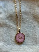 Collar dorado con cadena de eslabones pequeños y dije circular con diseño abstracto en tonos rosados y detalle de media luna dorada.