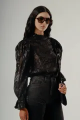 Blusa negra de manga larga confeccionada en tul semitransparente con bordado de lentejuelas en diseño abstracto. Presenta cuello alto fruncido y hombros ligeramente marcados con volumen.