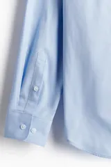Camisa celeste de algodón con cuello italiano, tapeta clásica y canesú en la espalda. Mangas largas con tapeta y botón, y cierre ajustable de botón en los puños. Bajo levemente redondeado. Corte ajustado que realza el contorno del cuerpo creando una silueta entallada.