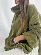 Buzo de pana fina color verde oliva, con diseño de corte holgado y mangas largas. Presenta una textura de cordón vertical característica de la pana y un pequeño parche decorativo en la parte superior de la espalda.