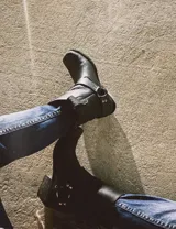 Bota corta de cuero color marrón, con taco de madera y punta cuadrada. Presenta una correa con hebilla alrededor del tobillo.