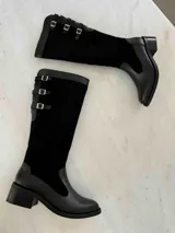 Bota alta de cuero negro con caña de gamuza negra y detalles de hebillas plateadas. Tiene un taco cuadrado bajo y cierre lateral. El diseño combina texturas y detalles metálicos para un estilo moderno y elegante.