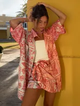 Falda pareo corta, color coral, con estampado de temática playera que incluye sellos postales, conchas marinas y texto. Se anuda en la cintura.