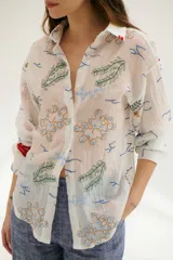 Camisa beige de manga larga con cuello clásico y cierre frontal con botones. Presenta bordados con motivos florales y geométricos en tonos azules, rojos y verdes.