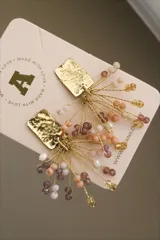 Par de aros colgantes con base rectangular dorada con textura martillada, de la que cuelgan hilos dorados con cuentas de color lila, blanco y naranja.