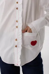 Camisa blanca de algodón con cuello alto con volados, cierre frontal con botones, mangas largas con puños abotonados, bolsillo en el pecho con corazón bordado y parches de crochet en los codos.