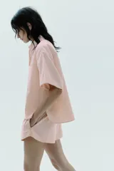 Camisa color rosa claro a rayas verticales finas color marrón, de popelín, con cuello solapa y manga corta con dobladillo. Tiene un bolsillo de parche en el pecho, aberturas laterales en el bajo y pinzas en la espalda. El cierre frontal es con botones ocultos.