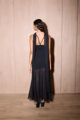 Vestido negro de largo midi, con tirantes finos y escote recto. La falda es asimétrica, con una capa superior de gasa transparente que cae hasta el tobillo y una base más corta de crepé elastizado. Incluye una bufanda larga de gasa a juego que se ata al cuello.