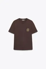 Camiseta marrón de algodón de manga corta y cuello redondo. Presenta un bolsillo en el pecho con un broche metálico dorado con diseño intrincado.