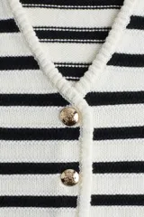 Chaleco corto de punto con rayas horizontales negras y blancas, escote en V y botonadura frontal con botones dorados. Ribetes acanalados en cuello, sisas y bajo.