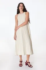 Vestido largo de tejido de punto fino con Lurex, color crema o beige claro. Presenta un diseño plisado vertical y cuello halter sin mangas, con silueta en A.