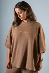 Remera color marrón claro de punto Prato, manga corta y calce holgado. Presenta bordado frontal con la palabra "bowwmana" en color naranja.
