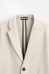 Abrigo recto color beige confeccionado en tejido con lana. Tiene cuello con solapas de muesca, manga larga con puños abotonados, bolsillos de vivo en la cadera, bolsillo interior, abertura en el centro de la espalda y cierre frontal con botonadura.