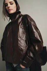 Chaqueta de Zara Woman Collection en tejido con acabado brillante efecto cuero. Presenta cuello solapa, manga larga con trabillas ajustables, bolsillos delanteros y cierre frontal con botones ocultos bajo solapa.
