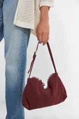 Cartera de hombro color bordó, confeccionada en gamuza, con correa ajustable y hebilla metálica.