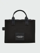 Bolso tote mediano de lona color beige con detalles en negro. Tiene doble asa de mano de cuero y correa bandolera ajustable y extraíble. Cuenta con dos bolsillos frontales con solapa y un bolsillo frontal con cierre. Estampado frontal con la leyenda "THE TOTE BAG" y el logo "MARC JACOBS".