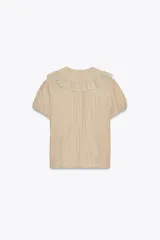 Camisa color crema con cuello bobo, manga corta abullonada con elástico y detalle de bordados combinados a tono y pliegues. Cierre frontal con botones forrados.