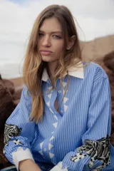 Camisa celeste a rayas verticales blancas, de corte holgado, con cuello clásico blanco, cierre frontal con botones y estampado de leopardo en los puños.