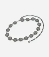 Cinturón de metal con acabado oscuro, compuesto por una cadena gruesa y una serie de dijes circulares con diseño floral en relieve. El cierre es de mosquetón con un dije floral como terminación.