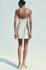 Vestido corto blanco de tejido elástico, con escote recto, tirantes finos ajustables, detalle de pliegues y cierre con cremallera oculta en la costura.