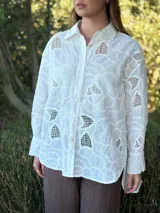 Camisa blanca de broderie con diseño floral, corte oversize, cuello clásico, cierre frontal con botones y mangas largas con puños abotonados.