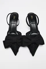 Zapato destalonado negro con tacón fino, capellada de mesh bordado y lazo decorativo en la parte delantera.