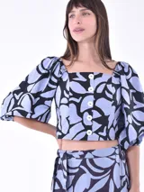 Blusa corta con estampado de hojas en tonos celeste y negro, escote cuadrado, mangas abullonadas 3/4 y cierre frontal con botones blancos.