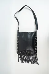 Cartera bandolera negra de cuero sintético con flecos en la parte inferior y detalles de ojalillos metálicos en los laterales.