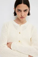 Cárdigan corto de punto texturizado color crema, con cuello redondo, cierre frontal con botones dorados y bolsillos frontales decorativos con botón dorado. Tiene terminaciones acanaladas en cuello, puños y ruedo.