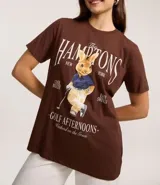 Remera de algodón color marrón oscuro con cuello redondo y manga corta. Presenta un estampado localizado con el texto "The Hamptons New York" y la ilustración de un conejo vestido de golfista.
