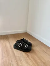 Sandalias marrones de cuero con doble tira y hebillas doradas, sobre base chunky negra.