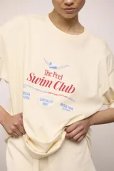 Remerón de manga corta color crema, corte oversize y cuello redondo. Estampado frontal con texto y un dibujo de una persona nadando, con la frase "The Peel Swim Club".
