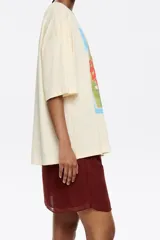 Camiseta color crema de algodón con estampado de un cuadro con borde celeste, con una mano sosteniendo una flor y el texto "Bimba y Lola".