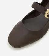 Zapatilla tipo bailarina de cuero color marrón, con punta redondeada y tira sobre el empeine con hebilla metálica dorada. Posee un tacón bajo de 1 cm y diseño clásico.