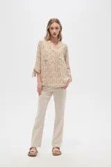 Blusa color beige con estampado de arabescos blancos, cuello mao, mangas 3/4 con lazos en los puños, cierre parcial con botones y bolsillos frontales.
